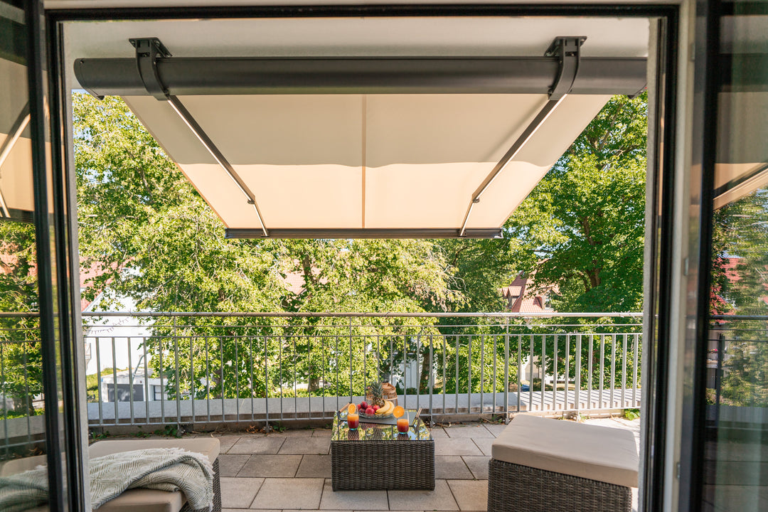 Tenda da sole color crema aperta sopra un balcone, vista dall'interno, con tavolino e poltroncine davanti agli alberi