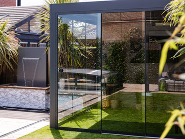 Pareti in vetro scorrevoli con telaio antracite su una pergola, con il prato e la piscina riflessi nei vetri