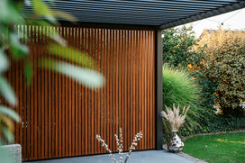 Parete a doghe in legno sotto una pergola a lamelle, con erba ornamentale e vaso decorativo in giardino