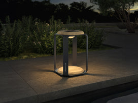 Lanterna solare Everglow antracite con base e coperchio in teak, accesa al crepuscolo su un bordo in pietra