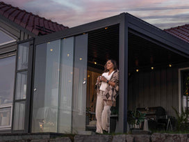 Donna con una tazza in mano davanti alle pareti in vetro di una pergola antracite, sul patio al tramonto