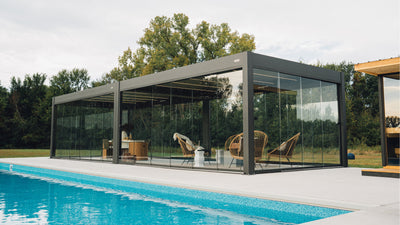 Pergola bioclimatica in alluminio scuro con pareti in vetro a bordo piscina e sedie in rattan all'interno