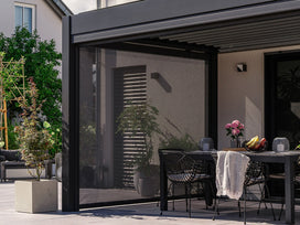 Tenda a rullo antracite con rete tessile abbassata a lato di una pergola, davanti a un tavolo da pranzo in terrazza